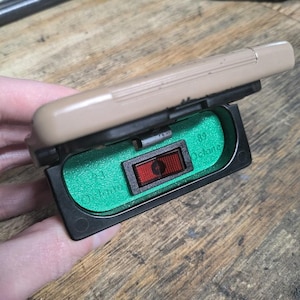 May include: A black and green plastic box with a red button labelled "On" and "Off". The box is open and the button is visible.