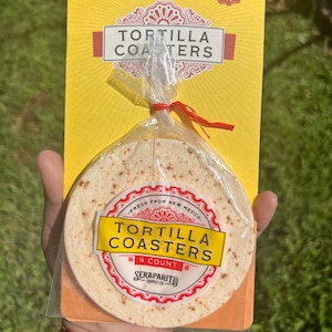 May include: A package of four tortilla coasters, made from fresh tortillas from New Mexico. The package is yellow with red and white text. The coasters are packaged in a clear plastic bag with a red ribbon.