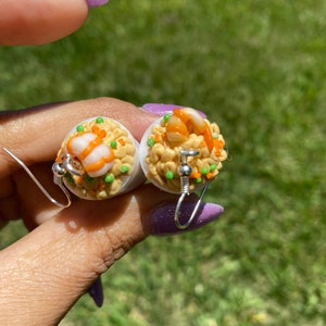 Puede incluir: Un par de pendientes con r&eacute;plicas de comida en miniatura de arroz frito con camarones. Los pendientes son blancos con detalles en naranja, amarillo y verde.