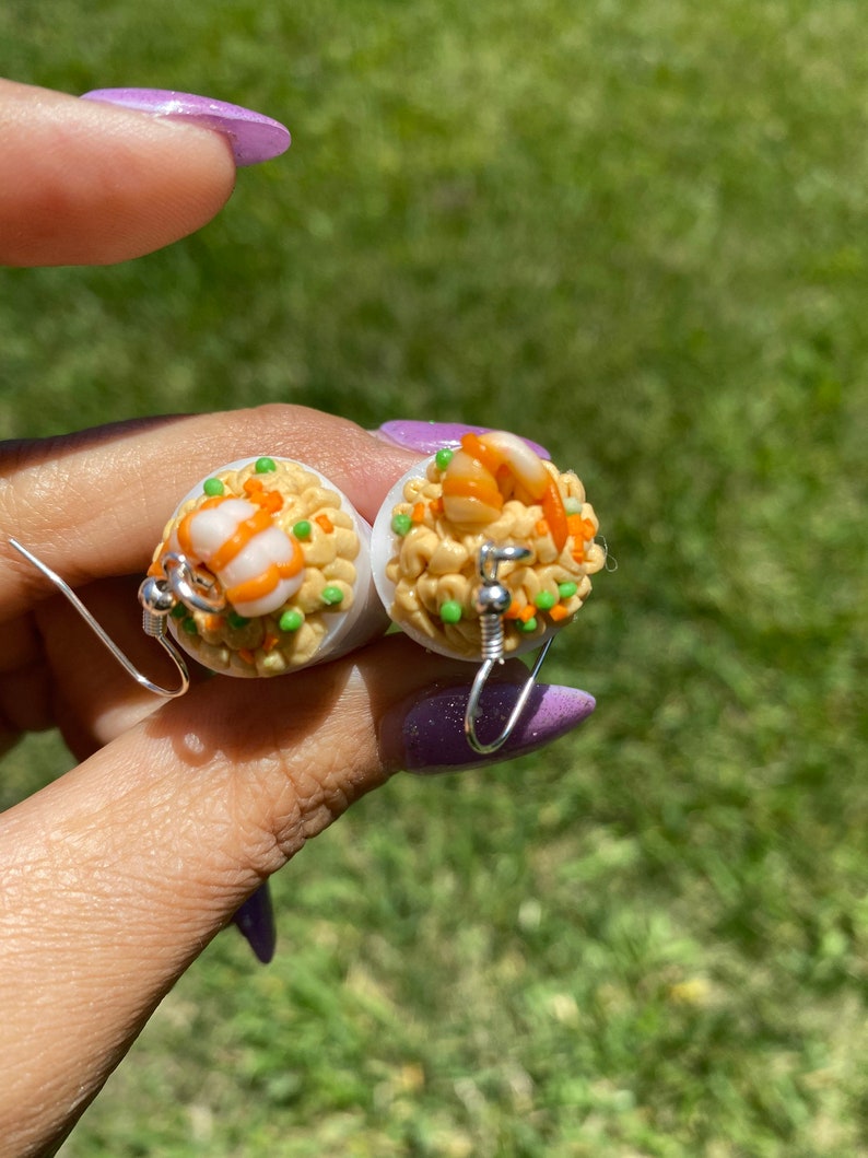 Puede incluir: Un par de pendientes con r&eacute;plicas de comida en miniatura de arroz frito con camarones. Los pendientes son blancos con detalles en naranja, amarillo y verde.