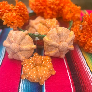 May include: Close-up of several pan de muerto, a traditional Mexican sweet bread, on a colorful striped textile. The bread is round with bone-shaped decorations and sugar coating. Orange marigold flowers and a green basket are in the background.