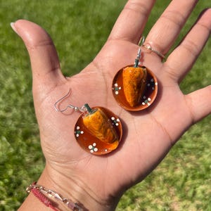 May include: A pair of handmade earrings featuring a miniature orange chili pepper design. The peppers are set on a clear, round base with a reddish-brown border and white and green accents. The earrings have silver-colored hooks.