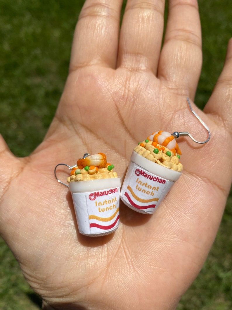 Puede incluir: Un par de pendientes con forma de tazas de ramen instant&aacute;neo. Las tazas son blancas con detalles rojos y amarillos y dicen "Maruchan Instant Lunch". Los fideos son amarillos y los ingredientes son guisantes verdes y camarones naranjas.