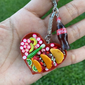 May include: A heart-shaped keychain with a miniature plate of tacos, limes, avocado, onions, and radishes. A miniature Coca-Cola bottle is attached to the keychain.