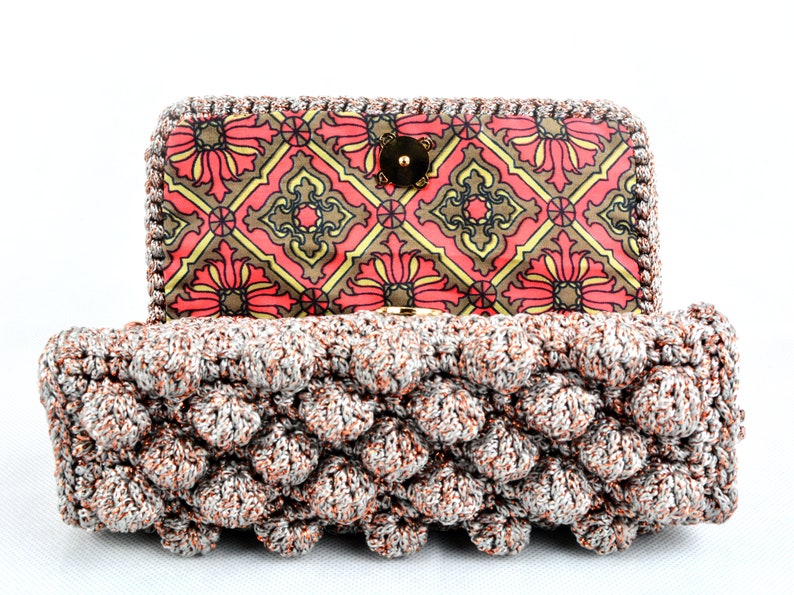 May include: A crocheted clutch purse with a silver and copper metallic yarn. The purse has a geometric patterned lining in shades of red, brown, and yellow.