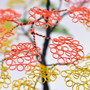 Pu&ograve; includere: Una scultura ad albero in filo di ferro fatta a mano con accenti arancioni, gialli e verdi. L'albero presenta intricati anelli di filo che formano le foglie, con un tronco e rami in filo attorcigliato nero e rosso. Lo sfondo &egrave; sfocato, evidenziando i dettagli colorati.