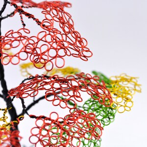 Pu&ograve; includere: Una scultura di albero in filo metallico fatta a mano con foglie rosse, gialle e verdi ad anello. I rami dell'albero sono neri e le foglie sono disposte a grappoli. Lo sfondo &egrave; bianco.