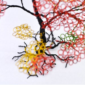 Pu&ograve; includere: Scultura ad albero in filo di ferro realizzata a mano con un tronco e rami attorcigliati neri. Il fogliame dell'albero presenta anelli rossi, verdi e gialli, creando un'esposizione vibrante e artistica. La scultura &egrave; su sfondo bianco.