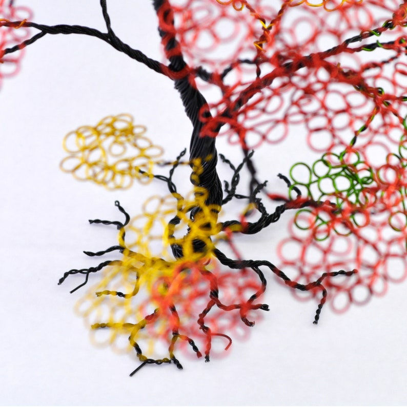 Pu&ograve; includere: Scultura ad albero in filo di ferro realizzata a mano con un tronco e rami attorcigliati neri. Il fogliame dell'albero presenta anelli rossi, verdi e gialli, creando un'esposizione vibrante e artistica. La scultura &egrave; su sfondo bianco.