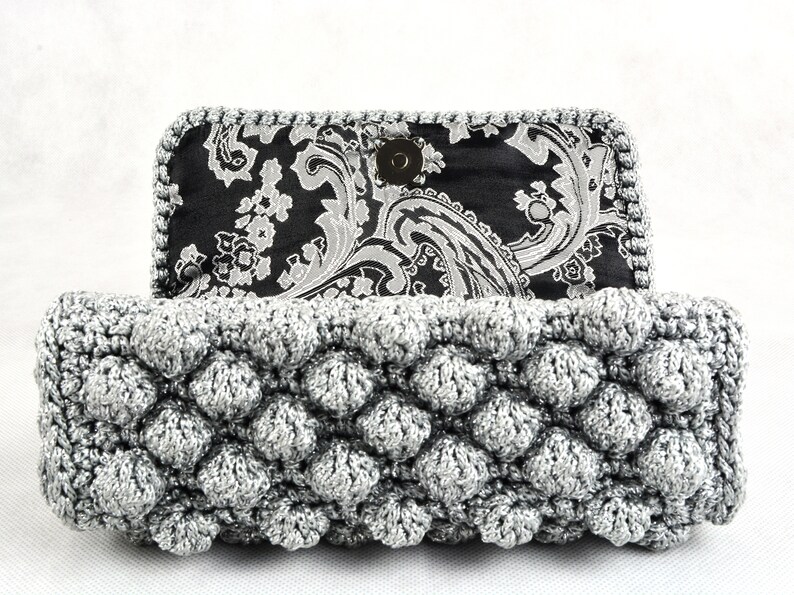 May include: A silver crocheted clutch purse with a black and white paisley fabric lining. The purse has a textured, bubble-like design.