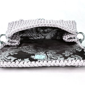 May include: A silver crocheted handbag with a black and silver paisley lining. The bag has a silver chain strap and a silver clasp.