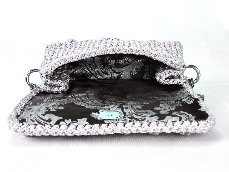 May include: A silver crocheted handbag with a black and silver paisley lining. The bag has a silver chain strap and a silver clasp.