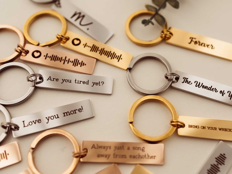 K&ouml;nnte beinhalten: Eine Sammlung von Schl&uuml;sselanh&auml;ngern aus Silber, Gold und Ros&eacute;gold mit gravierten Botschaften. Die Schl&uuml;sselanh&auml;nger zeigen verschiedene Botschaften, darunter "Forever", "The Wonder of You", "Are you tired yet?", "Love you more!", "Always just a song away from each other" und "Bring on your wreck".