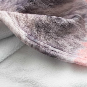 May include: A soft, plush throw blanket featuring a textured surface with a printed pattern of grey and black fur-like markings. The blanket has a solid light pink border along the edge and a plain white underside visible where the fabric folds.