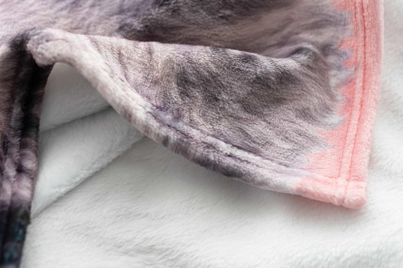 May include: A soft, plush throw blanket featuring a textured surface with a printed pattern of grey and black fur-like markings. The blanket has a solid light pink border along the edge and a plain white underside visible where the fabric folds.