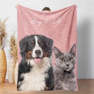 May include: A soft, dusty pink fleece blanket featuring a custom portrait of a Bernese Mountain Dog and a grey cat. The text "Zoey & Lola" is printed in white script at the top center of the blanket, positioned above the animal portraits.