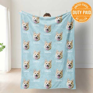Op de afbeelding: Een lichtblauwe fleece deken met een herhalend patroon van een hondenkop. De hond is een corgi met bruin en wit haar. De naam van de hond, "Shadow", staat in witte tekst op de deken.