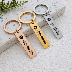 K&ouml;nnte beinhalten: Drei goldene und silberne Schl&uuml;sselanh&auml;nger mit einem einzigartigen Design. Jeder Schl&uuml;sselanh&auml;nger verf&uuml;gt &uuml;ber ein kleines Metallschild mit einem Barcode-&auml;hnlichen Muster und einem kleinen Symbol.