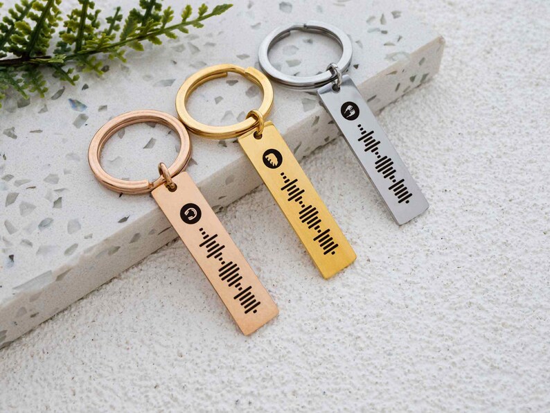K&ouml;nnte beinhalten: Drei goldene und silberne Schl&uuml;sselanh&auml;nger mit einem einzigartigen Design. Jeder Schl&uuml;sselanh&auml;nger verf&uuml;gt &uuml;ber ein kleines Metallschild mit einem Barcode-&auml;hnlichen Muster und einem kleinen Symbol.