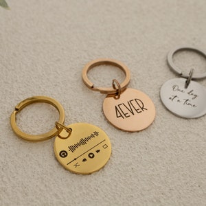 Könnte beinhalten: Drei Schlüsselanhänger in Gold, Roségold und Silber mit runden Metalletiketten. Der goldene Schlüsselanhänger hat ein Musiknoten- und Barcode-Design. Der roségoldene Schlüsselanhänger hat das Wort "4EVER" eingraviert. Der silberne Schlüsselanhänger hat den Text "One day at a time" eingraviert.