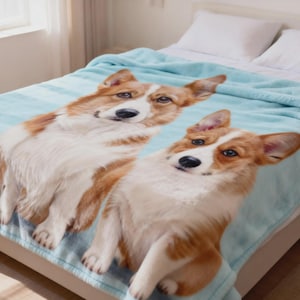 May include: A soft, light blue fleece blanket featuring a large, high-definition photographic print of two Corgi dogs. The dogs have tan and white fur, pointed ears, and dark eyes, and they are positioned side-by-side on the fabric surface.
