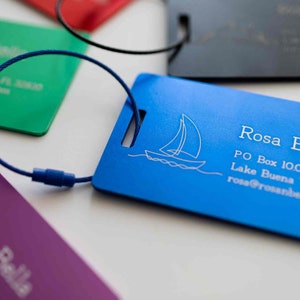 Könnte beinhalten: Ein blaues Gepäckanhänger mit einem weißen Segelboot-Design und dem Text "Rosa Bella", "PO Box 10000", "Lake Buena Vista, FL" und "rosa@rosabella.com".