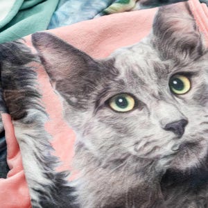 May include: A soft fleece blanket featuring a detailed graphic print of a grey cat with yellow-green eyes against a light pink background. The blanket has a textured surface and is folded over a sage green fabric with a visible stitched edge.