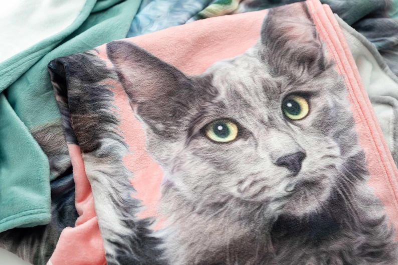 May include: A soft fleece blanket featuring a detailed graphic print of a grey cat with yellow-green eyes against a light pink background. The blanket has a textured surface and is folded over a sage green fabric with a visible stitched edge.