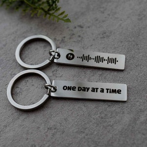 K&ouml;nnte beinhalten: Zwei silberne Schl&uuml;sselanh&auml;nger mit einem silbernen Ring. Ein Schl&uuml;sselanh&auml;nger hat einen Barcode und den Text "One day at a time".