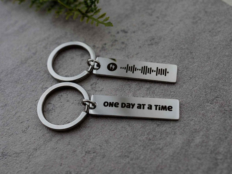 K&ouml;nnte beinhalten: Zwei silberne Schl&uuml;sselanh&auml;nger mit einem silbernen Ring. Ein Schl&uuml;sselanh&auml;nger hat einen Barcode und den Text "One day at a time".