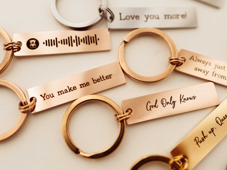 K&ouml;nnte beinhalten: Ein Satz Schl&uuml;sselanh&auml;nger aus Gold und Silber mit gravierten Botschaften. Die Botschaften lauten "You make me better", "Love you more", "Always just away from", "God Only Knows" und ein Barcode mit einem kleinen B&auml;ren-Symbol.