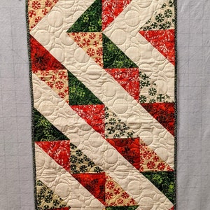 May include: A white table runner with a green and red geometric pattern. The pattern is made up of triangles and circles. The runner has a green and white border.