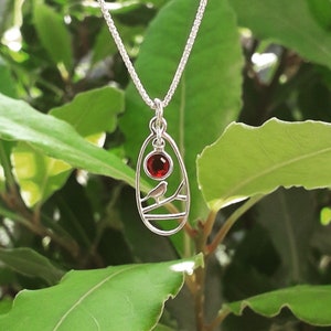 May include: A silver necklace with a pendant featuring a red gemstone and a bird on a branch. The pendant is shaped like an oval.