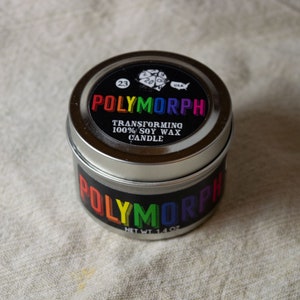 May include: A silver tin candle with a rainbow colored label that reads "POLYMORPH" and "TRANSFORMING 100% SOY WAX CANDLE". The label also includes a small image of a 20-sided die and the number "23".