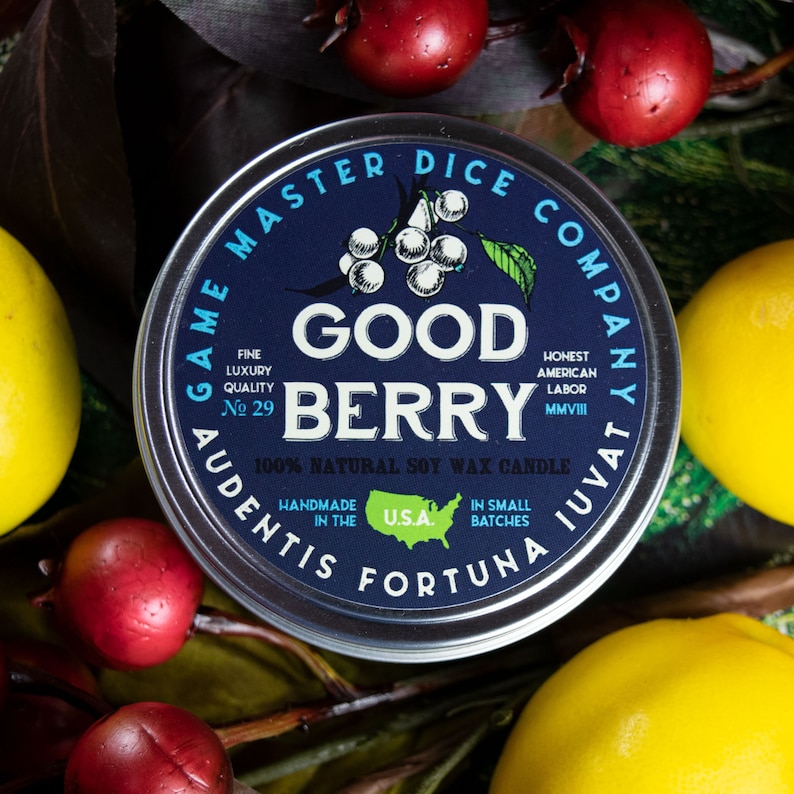 May include: A silver candle tin with a blue label that reads "Game Master Dice Company" and "Good Berry" in white lettering. The label also includes the text "100% Natural Soy Wax Candle" and "Handmade in the U.S.A."