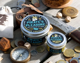 May include: An assortment of pirate-themed items, including candles, dice, a compass, coins, and a small leather pouch, arranged on a wooden surface. The candles are labeled "Pirate's Pleasure" and the compass has a brass finish.