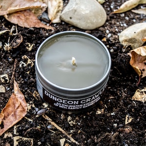 May include: A gray candle in a metal tin with the text "Dungeon Crawler" and "An excavating excursion of rich earthy garden soil".