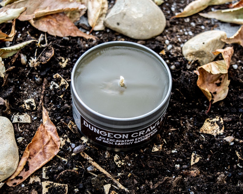 May include: A gray candle in a metal tin with the text "Dungeon Crawler" and "An excavating excursion of rich earthy garden soil".