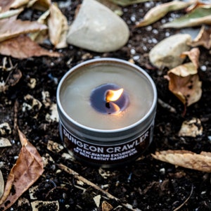 May include: A lit candle in a metal tin with the label "Dungeon Crawler: An Excavating Excursion of Rich Earthy Garden Soil". The candle is surrounded by brown leaves and rocks.