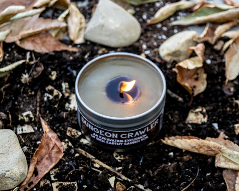 May include: A lit candle in a metal tin with the label "Dungeon Crawler: An Excavating Excursion of Rich Earthy Garden Soil". The candle is surrounded by brown leaves and rocks.