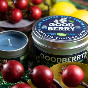 May include: Two candles in silver tins with blue and white wax. The larger tin has a blue label with the text "Good Berry" and "A Nourishing Lemon & Eucalyptus Scent". The smaller tin has a blue label with the text "Good Berry". Both candles are surrounded by red berries and lemons.
