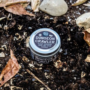 May include: A silver tin candle with a black label that reads "Dungeon Crawler" and "Clean-Burning 100% Soy Wax Candle". The candle is sitting on a bed of brown dirt and leaves.