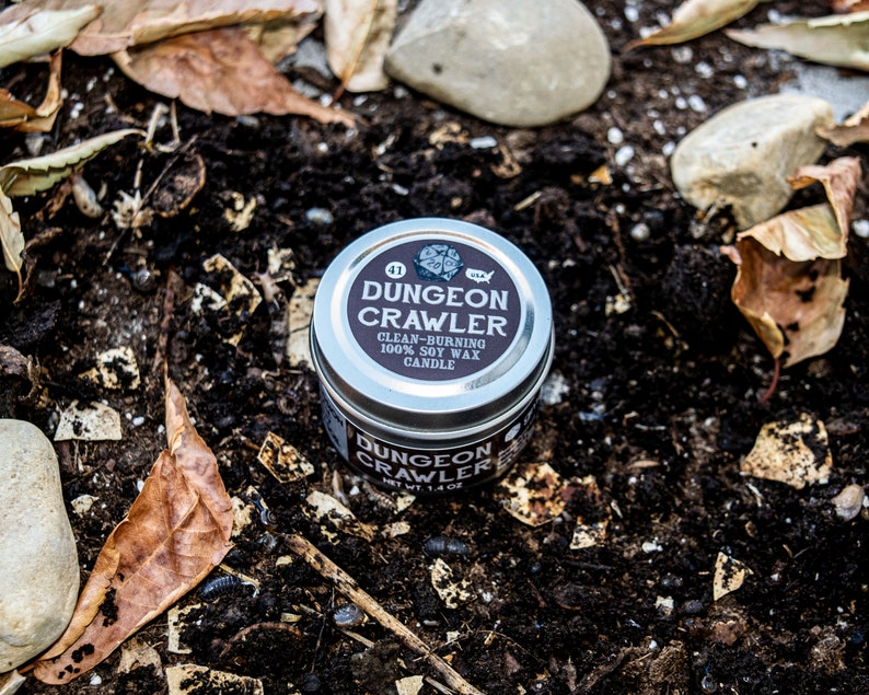 May include: A silver tin candle with a black label that reads "Dungeon Crawler" and "Clean-Burning 100% Soy Wax Candle". The candle is sitting on a bed of brown dirt and leaves.