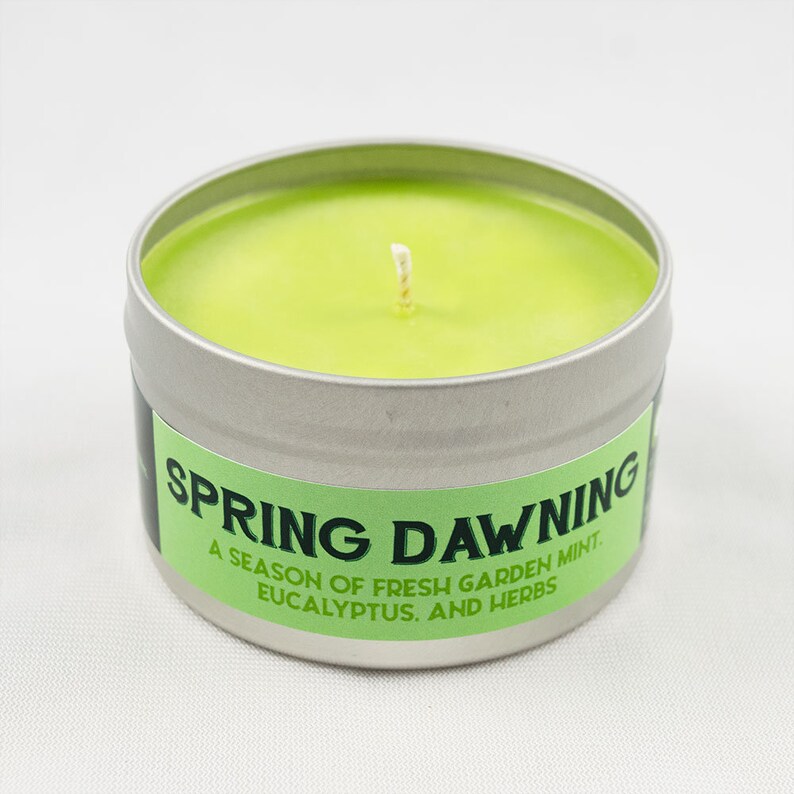 May include: A green candle in a silver tin with the text "Spring Dawning. A season of fresh garden mint, eucalyptus, and herbs."