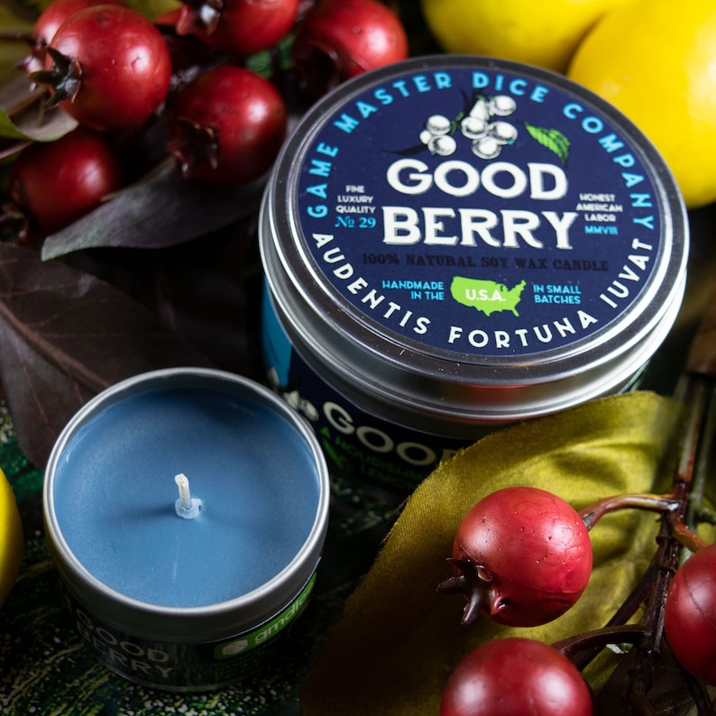 May include: A blue candle in a tin container with the label "Good Berry" and the text "Game Master Dice Company Audentis Fortuna Iuvat" in a circle around the label. The candle is surrounded by red berries and green leaves.