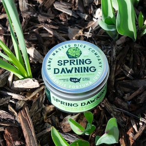 May include: A silver tin candle with a green label that reads "Spring Dawning" and "100% Natural Soy Wax Candle". The candle is surrounded by brown wood chips and green plants.