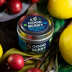 May include: A silver tin candle with a blue label that reads "Good Berry Clean-Burning 100% Soy Wax Candle" and "Good Berry Net Wt. 1.4 oz". The candle is surrounded by green leaves, red berries, and yellow lemons.