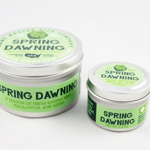 May include: Two silver tin candles with green labels. The larger candle is labeled "Spring Dawning" and says "A season of fresh garden mint, eucalyptus, and herbs". The smaller candle is labeled "Spring Dawning" and says "Clean-burning 100% Soy Wax Candle".