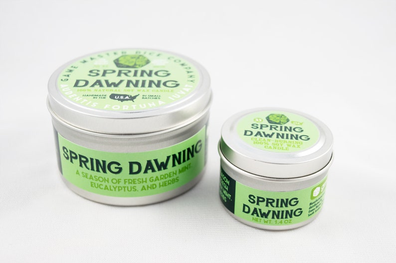 May include: Two silver tin candles with green labels. The larger candle is labeled "Spring Dawning" and says "A season of fresh garden mint, eucalyptus, and herbs". The smaller candle is labeled "Spring Dawning" and says "Clean-burning 100% Soy Wax Candle".
