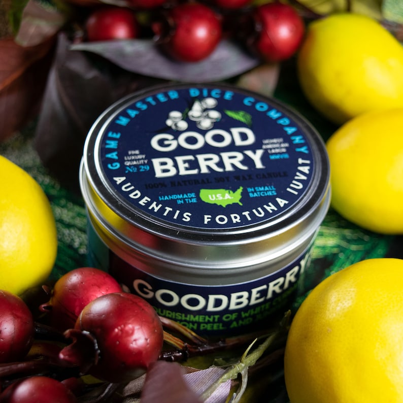 May include: A silver candle tin with a blue label that reads "Game Master Dice Company" and "Good Berry 100% Natural Soy Wax Candle". The label also includes the text "Audentis Fortuna Juvat" and "Handmade in the USA".